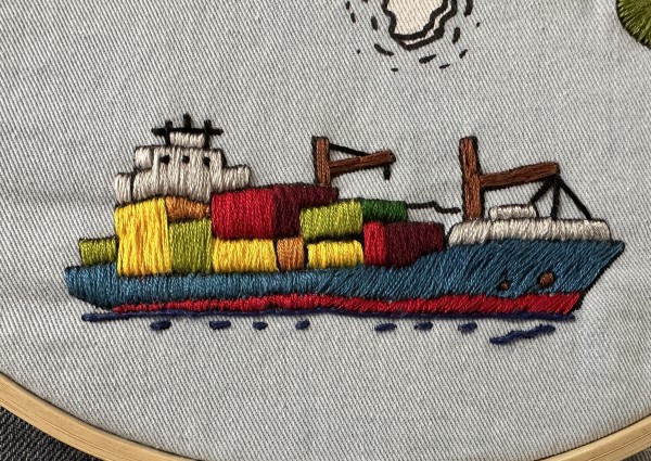 An embroidered cargo ship. The ship is blue with a white helm(?). There are lots of different coloured boxes or containers on the deck. 