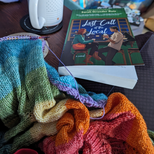 A copy of Last Call at the Local and a teacup sit beside an unfinished child sized rainbow sweater on knitting needles.