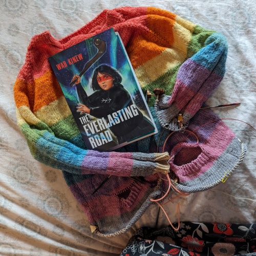 A unfinished kids sized rainbow striped sweater still on the knitting needles and a copy of the book The Everlasting Road by Wab Kinew. The book features a young indigenous woman on the cover holding a club with a background of Northern lights. 