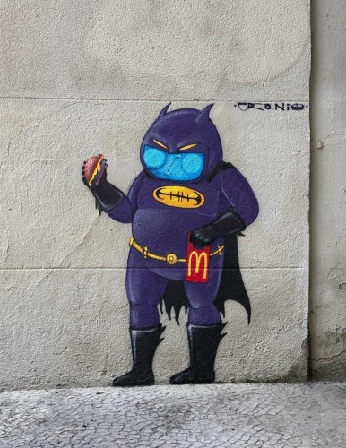 Streetartwall. A mural with a funny superhero in comic book style is sprayed/painted on a beige house wall. A Batman figure in a purple full-body suit and mask stands there with big cheeks and a hamburger in his hand. He has put on some weight because he is a bit chubby. In his other hand he is holding a snack bag from MC Donald's. The artist's name "Cranio" is written on the wall.