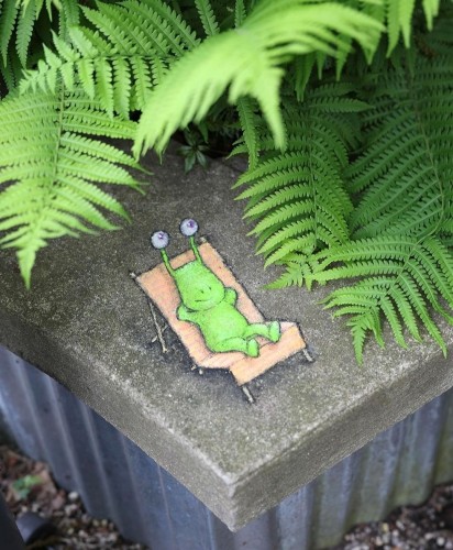 Streetart. A green creature (a snail without a shell) was painted with chalk on a low wall between green ferns. He (called Sluggo) is neon green, has stalk eyes, arms and legs. He is lying in a beige deckchair and enjoying the sun. Title : "Anyone who thinks it's not easy being green has never tried photosynthesis"