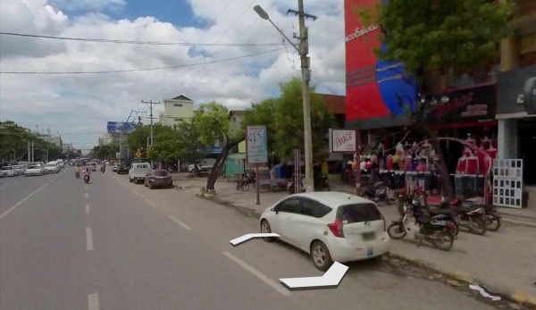 This image is a screenshot from Google Earth. It shows a busy street in Mandalay, Myanmar. Cars and small trucks zoom past under a cloudy sky. To the right is a red commercial building with shops displaying their wares on the ground floor. A sign outside this building says "Peace" in English, and I hope that the people there are indeed able to find peace, even as their military government persecutes them. 