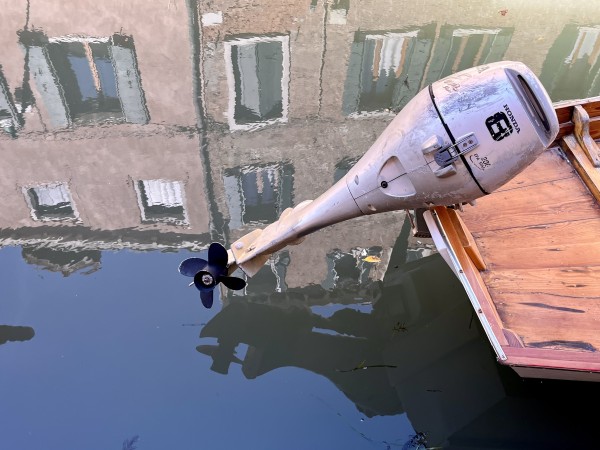 Colour photo of a canal, with, in the foreground, the wooden stern of a small boat, upon which is attached a scratched and slightly dented outboard engine with a tiny black propeller, looking for all the world like an immersion blender, raised out of the perfectly still and mirror-like water. The top half of the image shows the reflections of the building opposite, with two storeys' worth of windows with white marble surrounds and dark green shutters. 