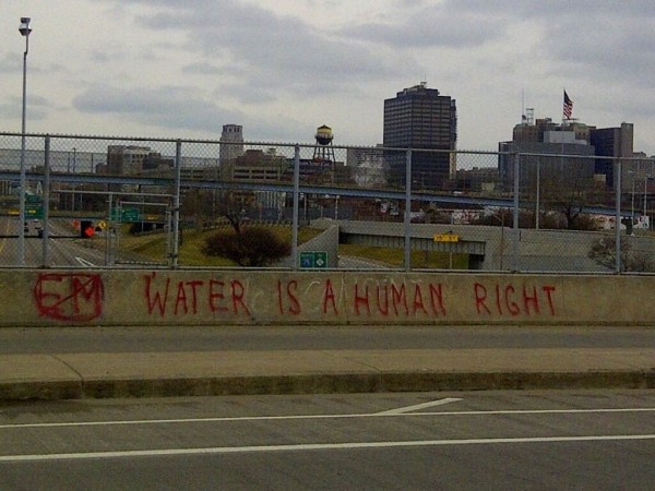 Pintada en un paso elevado de peatones sobre una autopista: "Water is a human right". Al fondo, rascacielos, un depósito elevado de agua, una bandera estadounidense.