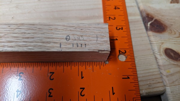 A block of wood nestled against a carpenter's square. The wood is marked at certain points along it's edge, and numbers are visible between the marks. 