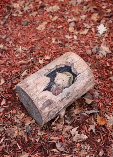 StreetArt. Auf ein kurzes Stück eines Baumstammes, der im Laub liegt, gezeichnet. Es zeigt einen kleinen Bär, der gemütlich im Baumstamm drinnen sitzt und ein Buch liest.