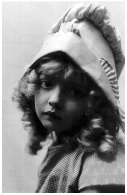 Black and white photo. Undated. We can see a girl with blondish curly hair. She is wearing a hood matching her dress.