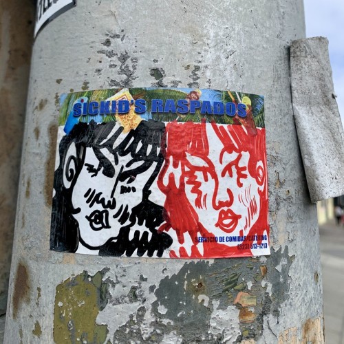 A graffiti slap sticker spotted on the street. It has a drawing of two women, with their faces both posed close to the viewer and filling up the frame. One is all black ink, the other is all red, they both look kind of mean.  The art is drawn in sharpie on a sticker label that is mostly white, but has "Sickid's Raspados" printed on the top.