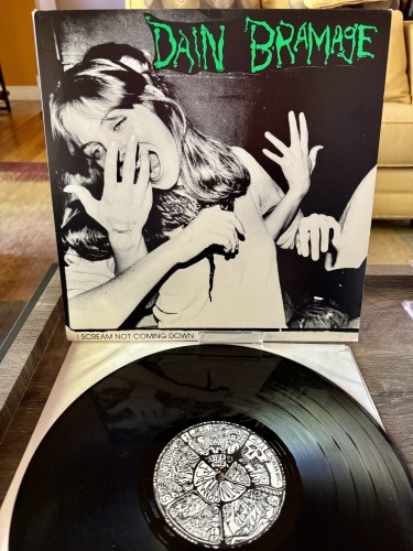 Front album cover. A female appearing person is in an awkward pose with another pair of hands coming from behind. She seems to be shouting at the hand. "Dain Bramage" spelled out in green macabre style text. 

Below is black vinyl disc with black and white label
