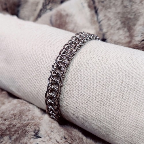 Bracelet en maille, dont le motif est plutôt plat et ressemble à un ruban. Les anneaux sont en acier inoxydable, donc non colorés. Le bracelet est disposé sur un support pour émuler un bras.