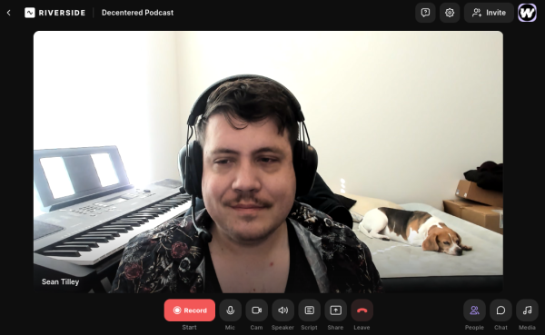 Sean, a white brown-haired man-boy with a moustache, wearing a flowery outer shirt over a sleeveless black shirt, wearing headphones and slightly smiling as a beagle snoozes on a bed behind him.