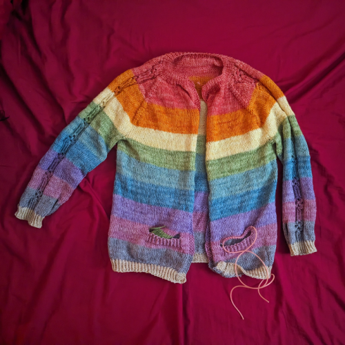 A nearly finished rainbow striped kid sweater in progress. One of the pockets still has a stitch holder with live stitches still on it. The sweater in an open-front raglan and features a faux cable motif on the sleeves. 