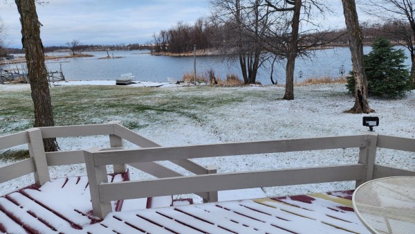 A lake sceen with a light dusting of snow on the green grass, and snow on a wooden deck overlooking the lake. The sky has light high level clouds. Bare trees line the shore.