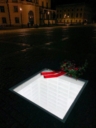 Photo of the "Empty Library" memorial at night. The window in the ground is illuminated from the room below. Someone put a flower wreath on top.