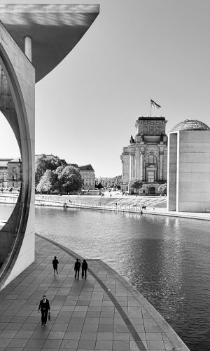 Black and white image showing people walking along a river Spree, framed by modern architecture, with former Reichstag- building and flag in the background.