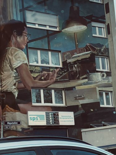 Eine Sekretärin mit Schreibmaschine und Kaffeetasse bei der Arbeit ist auf einer großen Hauswand aufgemalt. Zwischen ihren Arbeitsutensilien sieht man die Fenster des Gebäudes. 