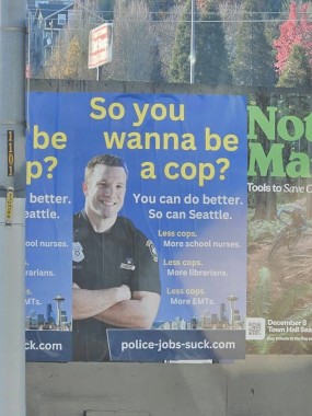 Picture of a poster with a smiling white cop with his arms folded. The Seattle skyline is in the background. The text reads "So you wanna be a cop? You can do better. So can Seattle. Less cops. More school nurses. Less cops. More librarians. Less cops. More EMTs. police-jobs-suck.com"