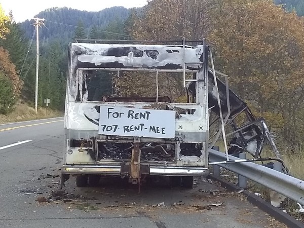 burned out shell of a travel trailer on the side of a forested mountain highway with a sign that reads:

"For Rent - 707-RENT-MEE"