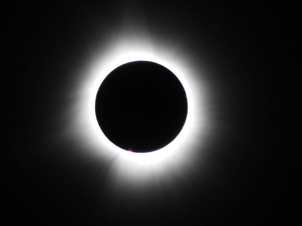 Close-up of a total solar eclipse, with a platinum colored ring and some orange and red solar flares visible around the edge