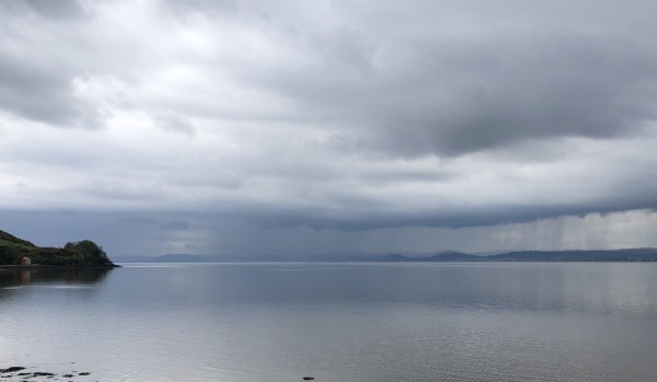 View over the Tay River looking upstream. The flat water is reflecting the colour of the grey clouds above. 
The clouds are pouring rain on the far shore while there are still some bright clouds above. A headland juts into view on the left hand side. 
