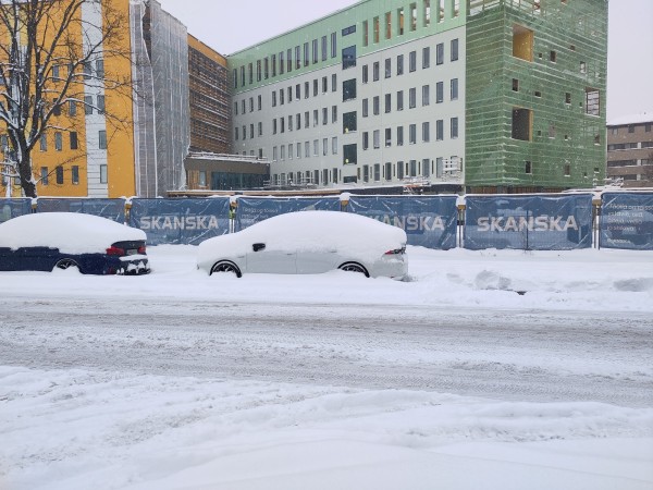 a picture taken from a snowy sidewalk. shows a road covered with snow. no asphalt is visible. two cars are parked on the side of the road covered with around 20 centimeters of snow. their tier rims are almost entirely covered by snow on the ground. In the background is a multistory tall building under construction but at last phases of construction. There is a tree visible at the left side of the picture that has no leaves and has snow on its branches.