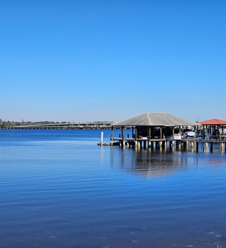 Overlooking a clear blue river beneath clear blue skies where a long bridge and adjacent pier are visible crossing the water. A number of large wood boat docks line the shoreline.