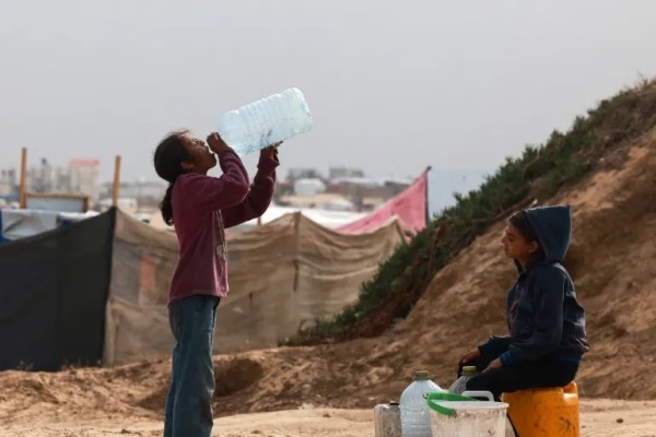 palestinian child drinking from a large bottle with Rafah tent city in the background