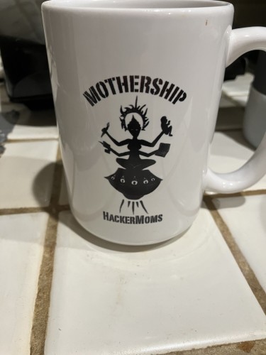 A mug w/ a divinely haloed multiarmed figure seated on a floating eye-ringed platform brandishes a paintbrush, powered drill, laptop computer & human baby. It says Mothership above her & HackerMoms below. 
