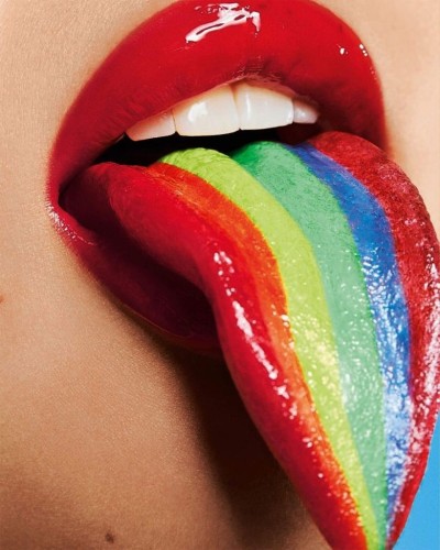 A close-up photo showing a woman wearing bright red lipstick with her tongue out. The woman's tongue is rainbow colored.