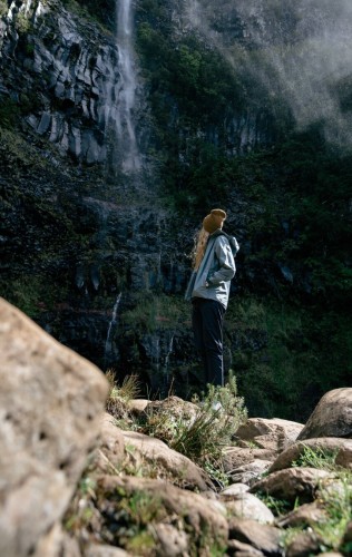 Photo of a woman looking at a waterfall, with stones in the foreground, waterfall, greenery and bushes in the background.