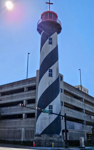 A favorite photo spot in downtown is showing its age under the scrutiny of the bright morning sun. A tall black and white striped lighthouse tower with a red top and Christian cross atop, connected to the multi level parking garage of a huge downtown church.