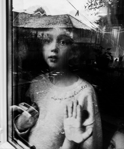 Photography. A black and white portrait photo of a little girl behind a window pane. The blonde child looks out of the window at the photographer with wide eyes. She is wearing a white woolen sweater with the words "allways in my heart" on the collar and a heart at the end of the lettering. To her right is a smaller boy, but you can only see his eye and his hand on the window pane. The neighboring houses are reflected in the window, giving the photo a melancholy and intangible atmosphere. A beautiful way to photograph a child's portrait.