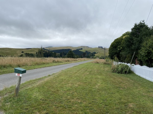 A rural roadside scene with a green mailbox numbered 82, a curving road, grassy fields, rolling hills in the background, and a cloudy sky. There's also a white picket fence and some foliage on the right side.