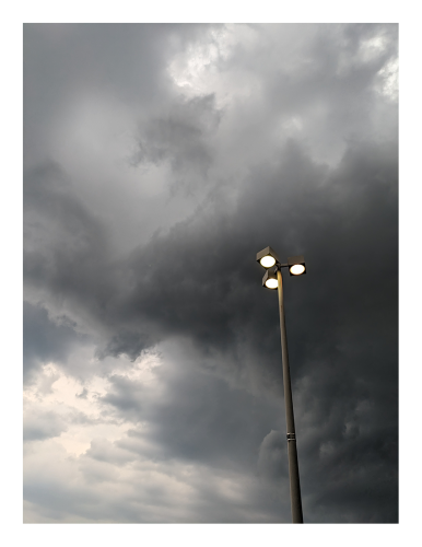 menacing gray and black clouds are forming in a section of the western sky on a late summer afternoon. a three-lamp street light rises to mid-picture at right.