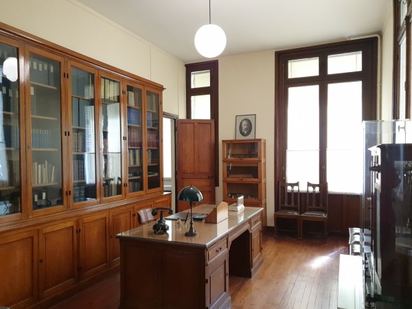 Photo of Marie Curie's former office. All dark wood, there's a big desk in the center. A cabinet with books behind it. On the other side,  large balance.
On a shelf beside the window in the back there's a photography of Pierre Curie (who was deceased at this point)