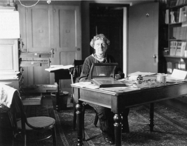 Annie Jump Cannon sat at a desk. She is clearly not a fan of a clear desk policy as it's messy with papers and open drawers. She is a white woman with pale hair.