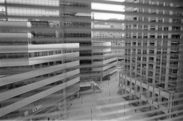 A black and white photograph through a Venetian blind of an empty modern city square. Virtually deserted, it looks more a model than real.