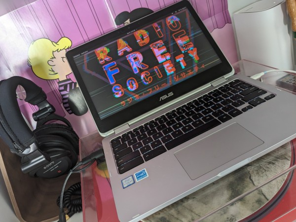 An Asus Chromebook laptop, running Pop!OS Linux with the screen open and on. The screen has a #RadioFreeSociety wallpaper displayed. The laptop sits on the clear lid of a red record player.