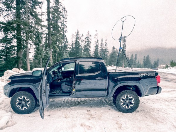 Snowy landscape with evergreen trees receeding into a mountainous backdrop. Black pickup truck with a strange looking looped antenna standing on a tripod and set on the bed of the truck.