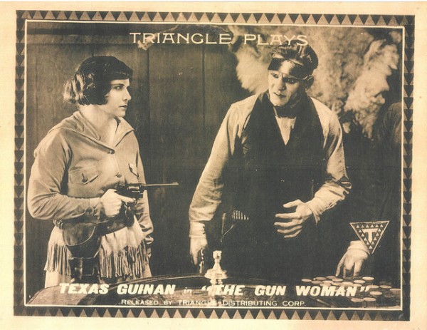 Black and white photo. A woman with dark, short, straight hair, wearing a long-sleeved dress is pointing a gun at a man. The man is wearing a long-sleeved white shirt and a black velvet. He is also holding a gun, but it is not pointed at the woman.