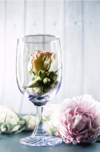 This is a still life photo of a glass cup with roses in it. A pink peony is next to it