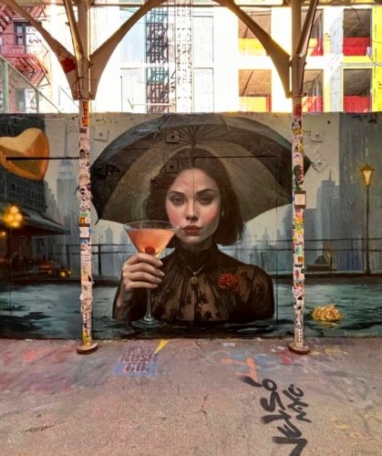 Streetartwall. A beautiful mural with a young, mysterious woman was sprayed/painted on a street wall. The lady with medium-length black hair is depicted up to her chest and is wearing a black lace dress, a rose as a pin and a golden heart on a chain. She is holding a cocktail glass with orange contents and a cocktail cherry. Above her is an old-fashioned green/grey umbrella. She is standing in the gray water and the silhouette of New York can be seen behind her. A pale rose floats by, a balloon flies beside her and a street lamp glows softly. A dark mural with a lot of atmosphere and a charismatic young lady.
