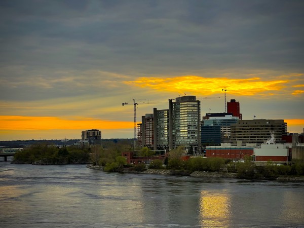 View of Gatineau from Ottawa shore. 
A large river with buildings and a sunset with god rays behind 