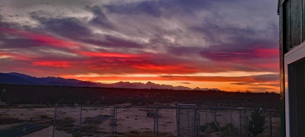 Streaky red and gold sunset across above a brushy, sandy desert. Mountains line the horizon. Chainlink panels are visible in the foreground, some assembled, some still stacked. The edge of a house intrudes on the right side of the picture.