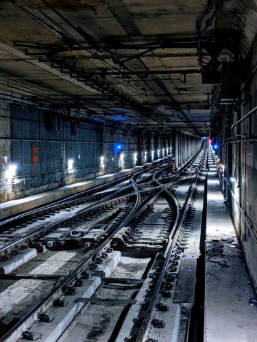 Concrete tunnel with 2 sets of train tracks going straight into the distance as well as a curved crossover in the middle. There are white and a few blue lights along the wall that illuminate the tunnel