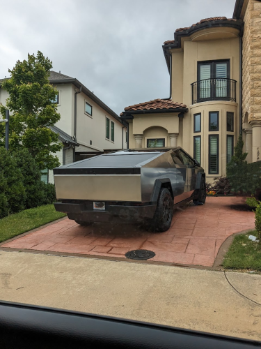 Photo of a new Tesla CyberTruck in Dallas. The truck is parked in a driveway of a house. Its brutalist lack of charm conflicts with the lovely Spanish colonial style home that it's parked at. 