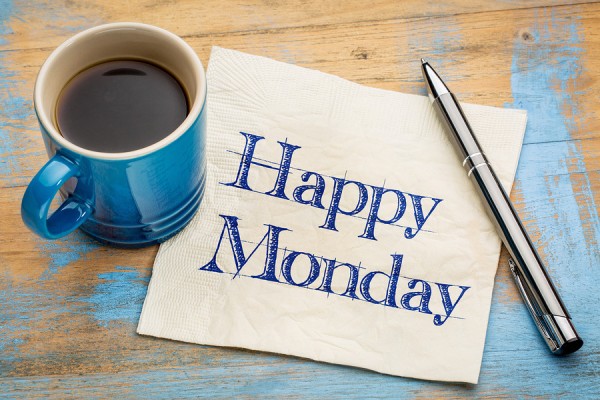 The picture shows a blue ceramic coffee mug filled with coffee on the left side. To the right of the mug, there is a white paper napkin with the phrase "Happy Monday" printed in blue, cursive font. On top of the napkin lies a silver pen with a black grip. The items are placed on a wooden surface with a rustic blue paint finish.