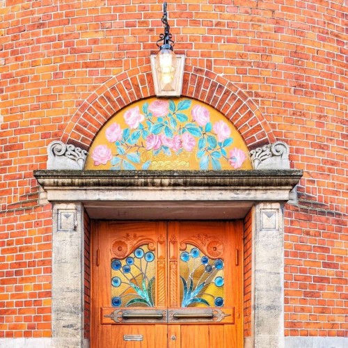 Top half of a wooden door with two stained glass panels. Instead of glass, the fanlight is painted with roses. There is a lamp over the door. The door is set against a round brick tower.
