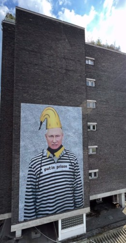 Streetartwall. A huge, satirical banner (10 x 6 meters) with a message about Vladimir Putin was placed on the outside wall of a six-storey modern building. It shows the Russian dictator in striped prison clothes with a yellow banana hat (like a pointed cap) on his head. Attached to his sweater is a sign that reads: "Put in prison". He looks grim. The background of the picture is a gray wall. 
Info: The artist Thomas Baumgärtel, alias Bananensprayer, is known for his ironic and satirical art campaigns and murals. His trademark is a yellow banana.