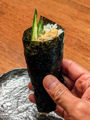 A cone of hanrolled sushi held up - some cucumber slices are sticking out at the wider open top of the handroll